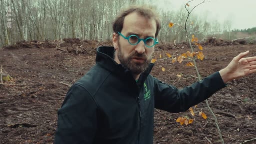 Un homme portant des lunettes à monture bleue et une veste noire s'exprime devant une parcelle de forêt déboisée, illustrant les conséquences écologiques de la gestion forestière.
