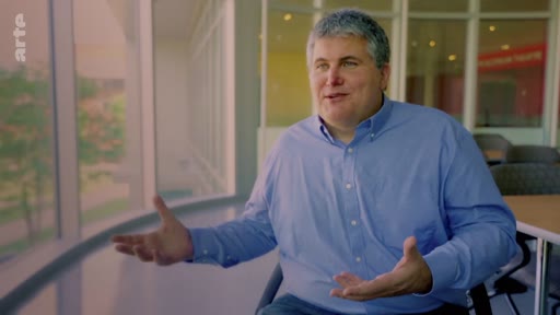 Un homme d'âge mûr, vêtu d'une chemise bleue, s'exprime avec les mains lors d'une interview filmée dans un espace de bureau lumineux avec de grandes baies vitrées. Il discute probablement des enjeux liés aux plantations industrielles d'arbres et à la foresterie.