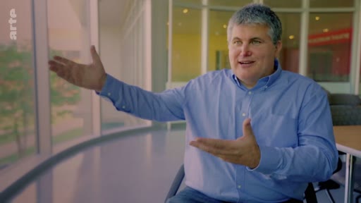 Un homme en chemise bleue s'exprime avec des gestes de la main lors d'une interview, probablement dans le cadre d'un reportage sur les plantations industrielles d'arbres.