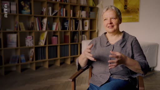 Une femme aux cheveux courts, assise dans un fauteuil, s'exprime lors d'une interview filmée devant une bibliothèque remplie de livres. Elle discute des risques écologiques liés à la monoculture dans le cadre d'un reportage pour la chaîne Arte.
