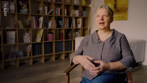 Une femme aux cheveux courts est assise dans un fauteuil devant une bibliothèque remplie de livres, lors d'une interview sur les risques écologiques de la monoculture.