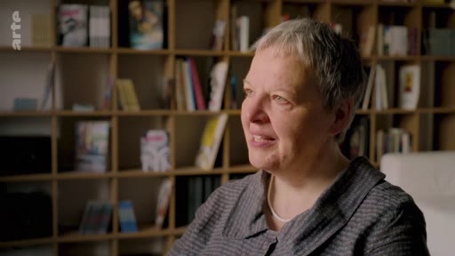 Une femme aux cheveux courts, vue en plan moyen, est interviewée devant une bibliothèque remplie de livres, abordant probablement des sujets liés à l'environnement ou à l'agriculture.