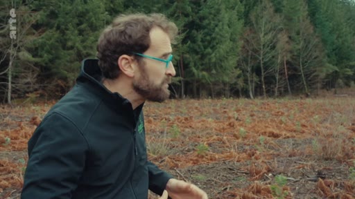 Un homme barbu avec des lunettes à monture bleue s'exprime lors d'un reportage sur l'opposition à la sylviculture industrielle, filmé en extérieur dans une zone forestière.