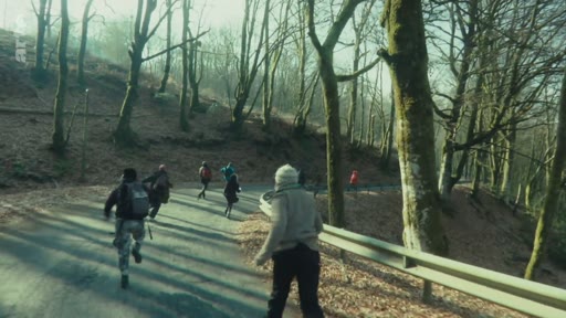 Des manifestants ou des personnes en fuite courent sur une route goudronnée traversant une zone boisée, identifiée comme le Bois du Chat. La scène montre une action de mouvement rapide dans un environnement naturel.