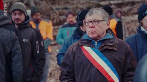 Un élu local portant l'écharpe tricolore française est entouré de professionnels de la filière bois lors d'un rassemblement en extérieur. La scène illustre une mobilisation ou une visite officielle dans le secteur forestier.