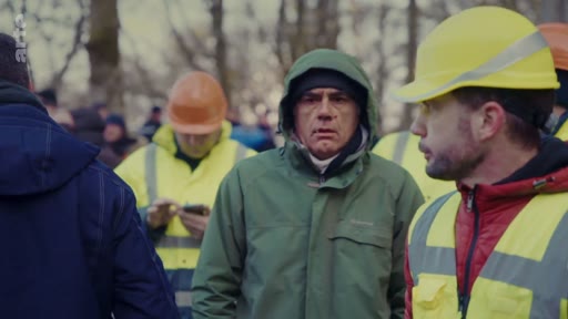 Un groupe de travailleurs forestiers, portant des casques de chantier et des gilets haute visibilité, est réuni en extérieur. L'un des hommes au premier plan porte un casque jaune et un gilet réfléchissant, tandis qu'un autre homme au centre porte une veste verte à capuche.