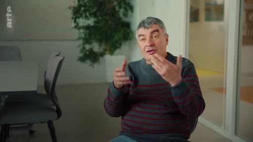 Un homme aux cheveux gris, vêtu d'un pull rayé, s'exprime lors d'une interview dans un cadre de bureau ou de salle de réunion. Il gesticule avec ses mains tout en parlant, illustrant une discussion sur des perspectives scientifiques ou durables.