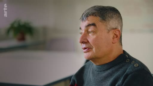 Un homme d'âge mûr aux cheveux gris est filmé en gros plan lors d'une interview, s'exprimant face à la caméra dans un environnement intérieur.