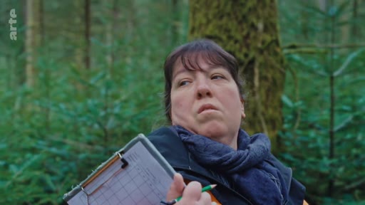 Une femme, probablement une forestière ou une scientifique, observe la régénération naturelle d'une forêt tout en notant des données sur un carnet. Elle porte une écharpe et une veste, travaillant en extérieur dans un environnement boisé.