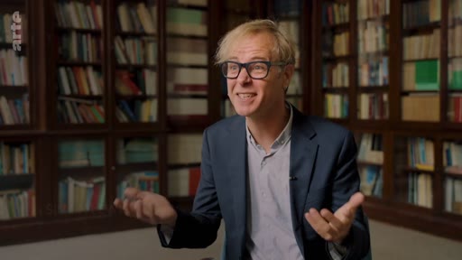 Un expert, portant des lunettes et une veste de costume, discute de la régénération naturelle des forêts lors d'une interview filmée dans une bibliothèque remplie de livres. Il utilise ses mains pour illustrer ses propos sur les avantages écologiques de ce processus forestier.