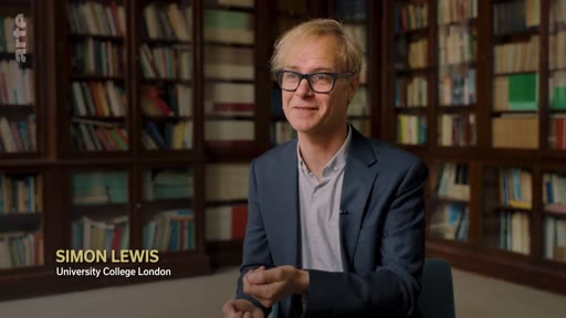 Simon Lewis, chercheur à l'University College London, s'exprime lors d'une interview sur le thème de la régénération forestière naturelle. Il est filmé en plan moyen devant des étagères remplies de livres.