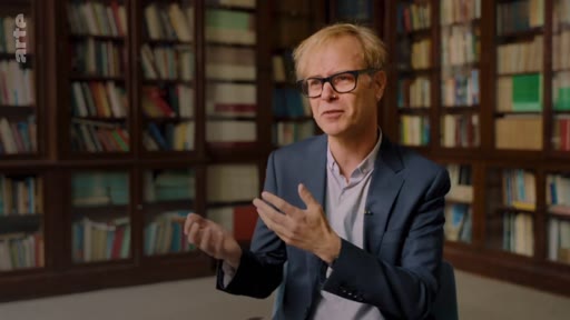 Un homme aux cheveux blonds portant des lunettes s'exprime lors d'une interview, avec une bibliothèque en arrière-plan. Il discute probablement des enjeux liés à l'expansion des plantations industrielles.