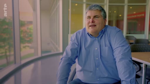 Un homme d'âge mûr, vêtu d'une chemise bleue, est filmé en plan moyen lors d'une interview dans un espace de bureau lumineux avec de grandes baies vitrées. Il s'exprime sur le thème de l'expansion mondiale des plantations industrielles.