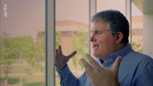 Un homme s'exprime avec les mains lors d'une interview filmée devant une grande baie vitrée, dans le cadre d'un reportage sur l'expansion industrielle des plantations.