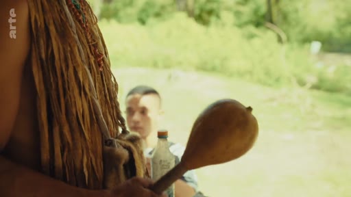 Un membre de la communauté indigène Pataxó, portant des ornements traditionnels, tient une maraca en bois lors d'un échange avec un jeune homme dans le territoire Pataxó au Brésil. Cette scène illustre la transmission culturelle et la vie quotidienne au sein de la communauté.