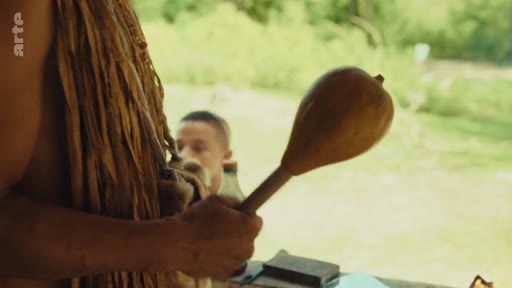 Un membre de la communauté autochtone Pataxó tient une maraca traditionnelle lors d'une cérémonie ou d'un rituel. En arrière-plan, un autre homme est visible dans un environnement extérieur, illustrant la culture et les traditions des peuples autochtones au Brésil.