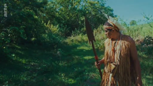 Un homme indigène vêtu d'une tenue traditionnelle en fibres végétales et portant une coiffe de plumes marche dans une zone boisée en tenant une lance. Cette image illustre la présence et le lien des peuples autochtones avec leur environnement naturel menacé.