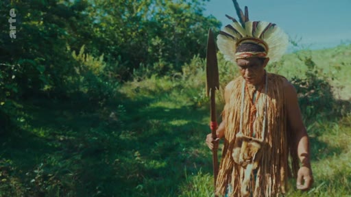 Un homme autochtone portant une coiffe en plumes et des vêtements traditionnels en fibres végétales marche dans une zone boisée en tenant une lance. Cette image illustre la culture et le mode de vie des peuples autochtones dans le contexte de la préservation de la forêt.
