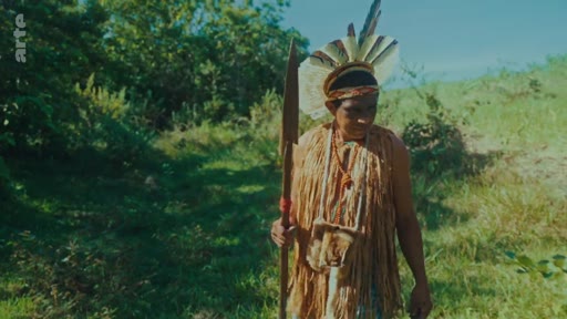 Un homme autochtone portant une coiffe en plumes et des vêtements en fibres naturelles marche dans un environnement forestier en tenant une lance. Cette image illustre la culture et le mode de vie des peuples autochtones face aux enjeux de la déforestation.