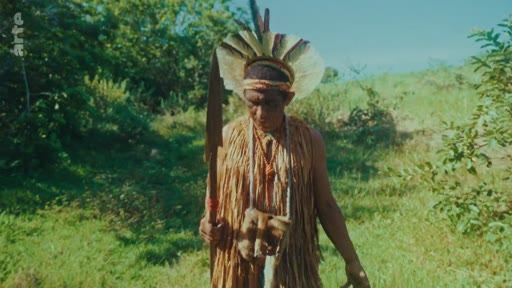 Un homme autochtone portant une coiffe en plumes et des vêtements en fibres végétales tient une lance tout en marchant dans un environnement forestier. Cette image illustre la culture et le mode de vie des peuples autochtones dans le contexte de la préservation de la forêt.