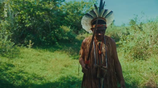 Un homme autochtone portant une coiffe traditionnelle et des vêtements en fibres naturelles marche à travers une zone boisée, illustrant la perspective des peuples indigènes sur la déforestation et la préservation de leur environnement.
