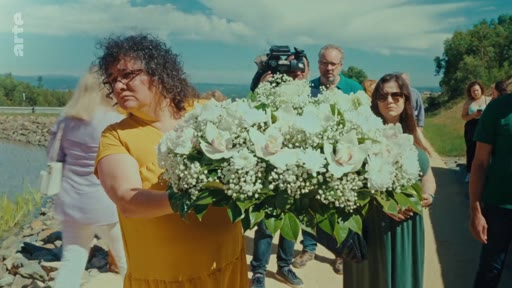 Des participants, dont une femme en robe jaune, portent une grande gerbe de fleurs blanches lors d'une cérémonie de commémoration en hommage aux victimes des incendies de 2017.
