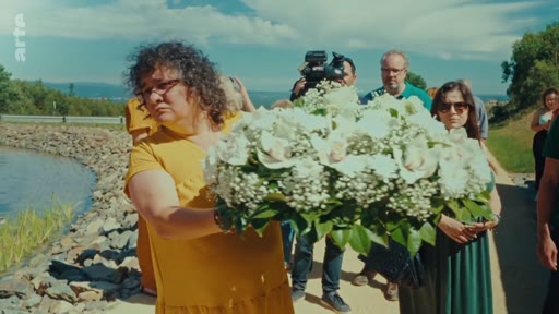 Une femme en robe jaune dépose une couronne de fleurs blanches lors d'une cérémonie en hommage aux victimes des incendies de 2017. La scène se déroule en extérieur près d'un point d'eau, en présence de plusieurs personnes.