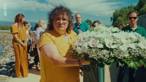 Une femme en robe jaune dépose une gerbe de fleurs blanches lors d'une cérémonie en extérieur, probablement en hommage aux victimes des incendies de 2017. Plusieurs personnes sont présentes en arrière-plan dans un cadre commémoratif.