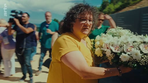 Une femme en haut jaune dépose une gerbe de fleurs blanches lors d'une cérémonie en extérieur, entourée de journalistes et de caméramans, dans le cadre de la commémoration des incendies de 2017.