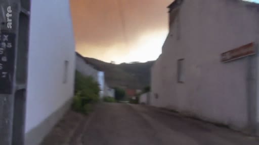Vue d'une rue étroite dans un village français plongé dans une atmosphère sombre et orangée, causée par la fumée d'un incendie de forêt à proximité. L'image témoigne de l'ambiance apocalyptique lors d'un feu de forêt majeur.