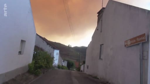 Vue d'une rue étroite dans un village portugais, avec un ciel teinté d'orange par la fumée d'un incendie de forêt. Un panneau de signalisation indique 'Praia Fluvial', illustrant la menace des feux de forêt sur les zones rurales.