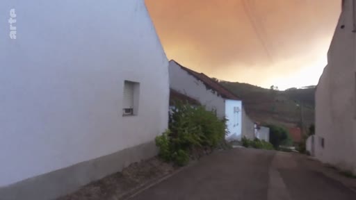 Vue d'une rue déserte dans un village, avec un ciel teinté d'orange par la fumée d'un incendie de forêt en arrière-plan. Cette image d'amateur illustre l'atmosphère apocalyptique causée par les feux de forêt.
