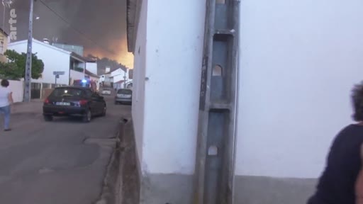 Des habitants évacuent une rue étroite alors qu'un incendie majeur fait rage à l'arrière-plan, avec une épaisse fumée noire visible au-dessus des maisons.