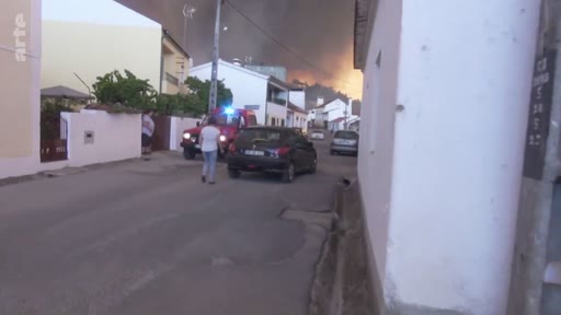 Des habitants observent un incendie de forêt menaçant une zone résidentielle, avec un véhicule de secours présent sur les lieux. L'image montre la tension lors d'une évacuation ou d'une intervention d'urgence face à un feu de forêt.