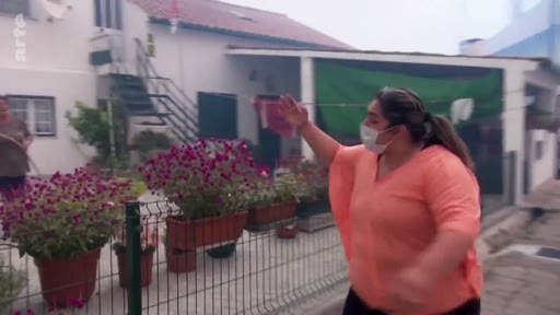 Une femme portant un masque de protection fait un signe de la main devant sa maison lors d'une évacuation liée à des incendies de forêt. Elle se tient devant une clôture avec des pots de fleurs dans un quartier résidentiel.