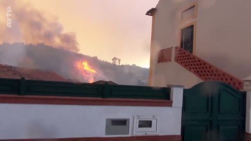 Vue d'une maison située à proximité d'un incendie de forêt en cours, avec des flammes et de la fumée visibles sur la colline en arrière-plan. L'image illustre la menace directe des feux de forêt sur les zones résidentielles.