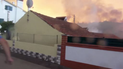 Vue extérieure d'une maison située à proximité immédiate d'un incendie de forêt, avec une épaisse fumée visible à l'arrière-plan. L'image illustre la menace des feux de forêt sur les zones résidentielles lors d'une évacuation.