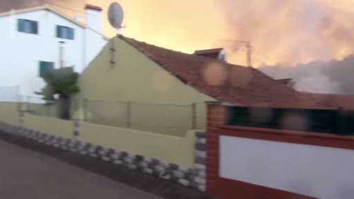 Une maison résidentielle située à proximité immédiate d'un incendie de forêt, avec une épaisse fumée orange visible à l'arrière-plan. L'image illustre la menace des feux de forêt sur les zones habitées.