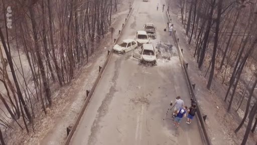 Vue aérienne d'une équipe de tournage filmant les restes de voitures calcinées sur une route après un incendie de forêt. La scène illustre les conséquences tragiques d'un sinistre naturel sur les infrastructures routières.