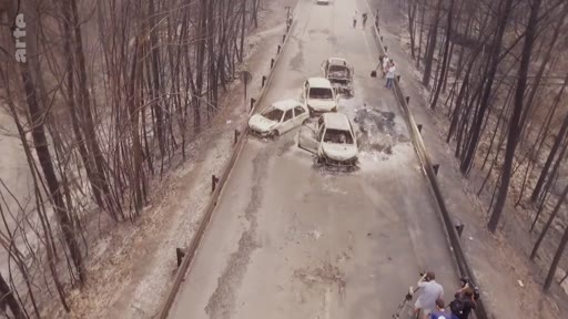 Vue aérienne montrant les conséquences d'un incendie de forêt sur une route où plusieurs véhicules ont été abandonnés et calcinés. Des personnes sont présentes sur les lieux pour constater les dégâts dans cette zone sinistrée.