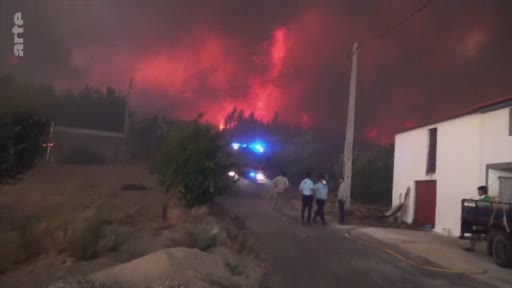 Des pompiers et des habitants observent un violent incendie de forêt qui illumine le ciel nocturne en rouge, menaçant des zones résidentielles. La scène montre l'urgence de la situation avec des véhicules de secours aux gyrophares allumés.