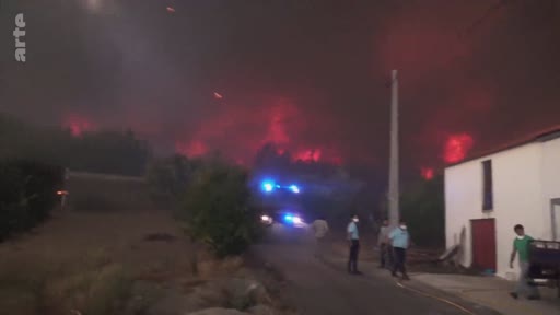 Des pompiers et des habitants observent un violent incendie de forêt nocturne, marqué par des flammes intenses et une fumée rougeoyante. La scène montre l'intervention des secours avec un véhicule équipé de gyrophares bleus dans une zone rurale.