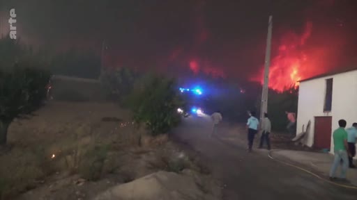 Des habitants et des intervenants observent un important incendie de forêt qui illumine le ciel nocturne en rouge, avec un véhicule d'urgence aux gyrophares bleus visible sur une route de campagne.