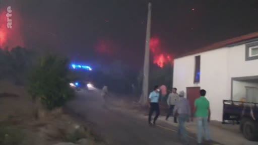 Des habitants se tiennent près d'une maison alors qu'un incendie de forêt illumine le ciel nocturne en arrière-plan. Un véhicule de secours avec des gyrophares bleus est visible sur la route à proximité, illustrant la lutte contre les feux de forêt.
