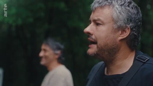 Un homme barbu aux cheveux gris s'exprime face caméra lors d'un entretien en extérieur, avec un autre homme flou en arrière-plan. Cette séquence s'inscrit dans le cadre de l'introduction du mouvement d'éradication des eucalyptus.