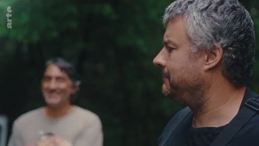 Un homme aux cheveux gris s'exprime lors d'un entretien en extérieur, avec un autre homme flou en arrière-plan, dans le cadre d'un reportage sur l'éradication de l'eucalyptus.