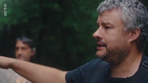 Un homme barbu aux cheveux gris s'exprime en extérieur, probablement dans le cadre d'un reportage sur l'éradication de l'eucalyptus. En arrière-plan, une autre personne est visible, floue.