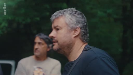 Un homme aux cheveux gris s'exprime lors d'une discussion en extérieur, probablement dans le cadre d'un reportage sur l'éradication des eucalyptus. Un autre homme est visible en arrière-plan.