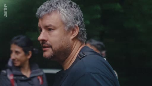 Un homme aux cheveux gris et à la barbe courte, participant au mouvement d'éradication de l'eucalyptus, s'exprime lors d'une séquence en extérieur. Il est accompagné d'une femme en arrière-plan dans un environnement boisé.
