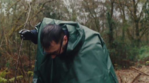 Un homme portant des lunettes et une cape de pluie verte s'occupe de la plantation d'un jeune arbre dans une zone boisée, illustrant des actions de reforestation et de préservation de l'environnement.